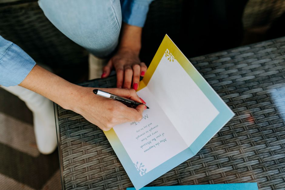 Femme écrivant dans une carte colorée sur une table en osier, avec un stylo noir et des ongles vernis.