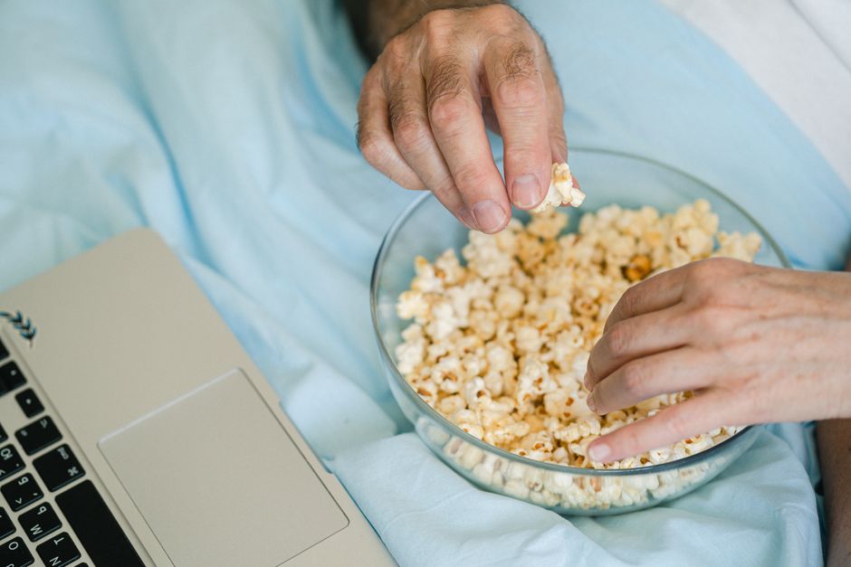 Mains tenant un bol de popcorn sur un lit, avec un ordinateur portable à côté, ambiance cosy pour une soirée film.
