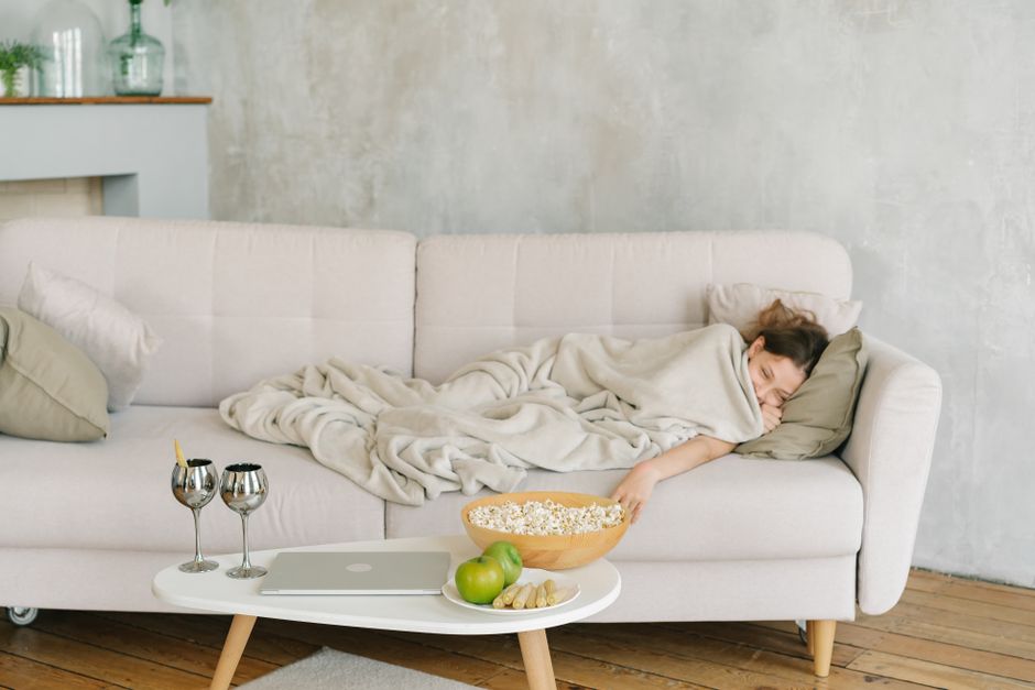 Femme emmitouflée dans une couverture sur un canapé, avec popcorn et boissons, prête pour une soirée film cosy.
