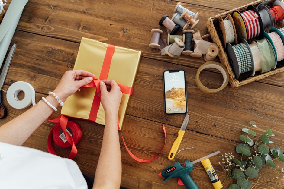 Personne en train d'emballer un cadeau avec un ruban rouge, téléphone et fournitures de bricolage sur une table en bois.