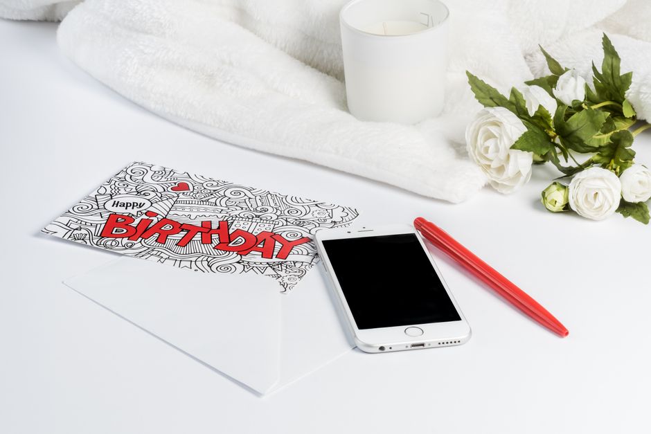 Carte d'anniversaire colorée avec un stylo rouge, un smartphone et des fleurs sur une surface blanche.