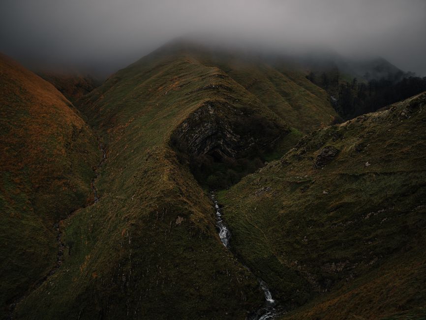 Paysage montagneux brumeux avec des collines verdoyantes et un ruisseau serpentant au fond de la vallée.