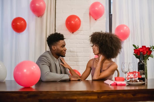Un couple romantique assis à une table décorée de ballons roses, échangeant des regards complices.