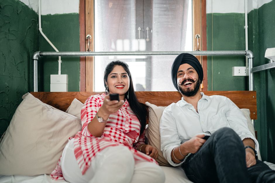Un couple souriant assis sur un lit, regardant la télévision avec une télécommande à la main.