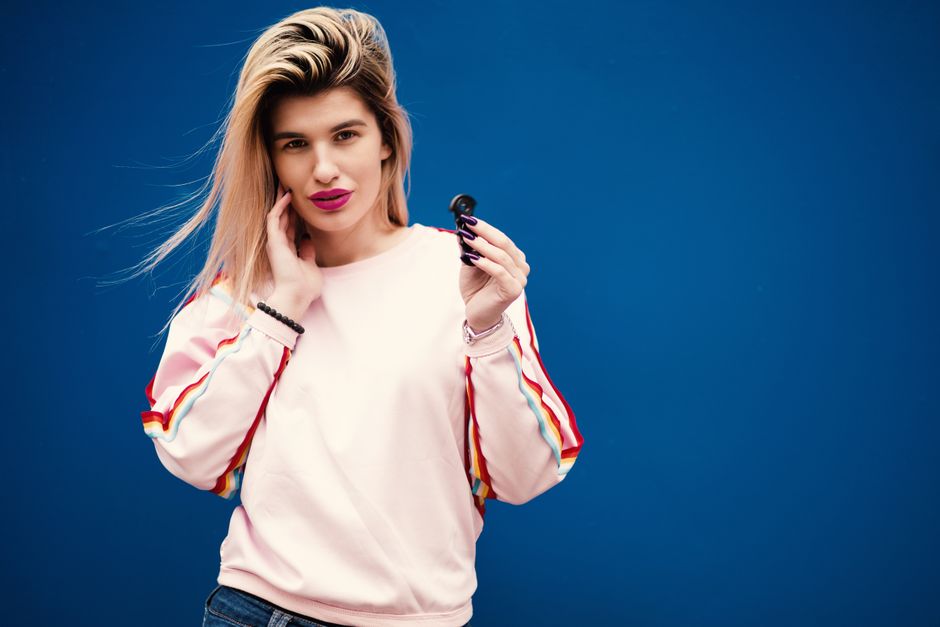 Femme souriante en sweat rose tenant un écouteur, devant un mur bleu, symbolisant la connexion dans le couple.