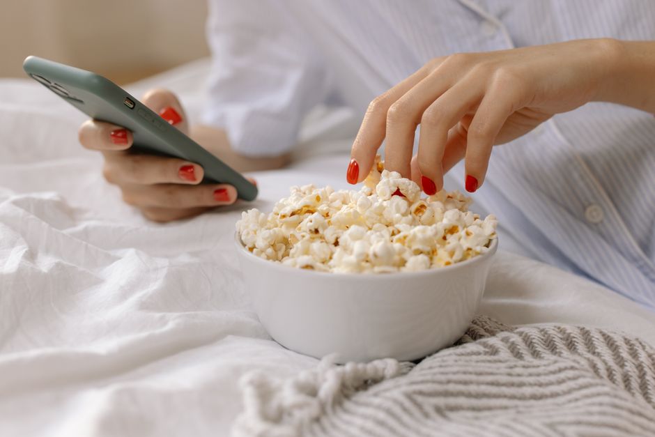 Main féminine avec vernis rouge prenant du popcorn dans un bol, téléphone à l'autre main, sur un lit cosy.