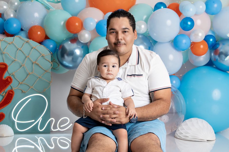Homme assis tenant un enfant dans un décor coloré de ballons pour une séance photo festive.