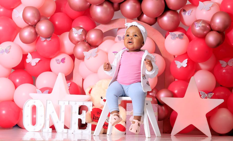 Un bébé assis sur une chaise devant un mur de ballons roses et rouges, avec des étoiles et un ours en peluche.