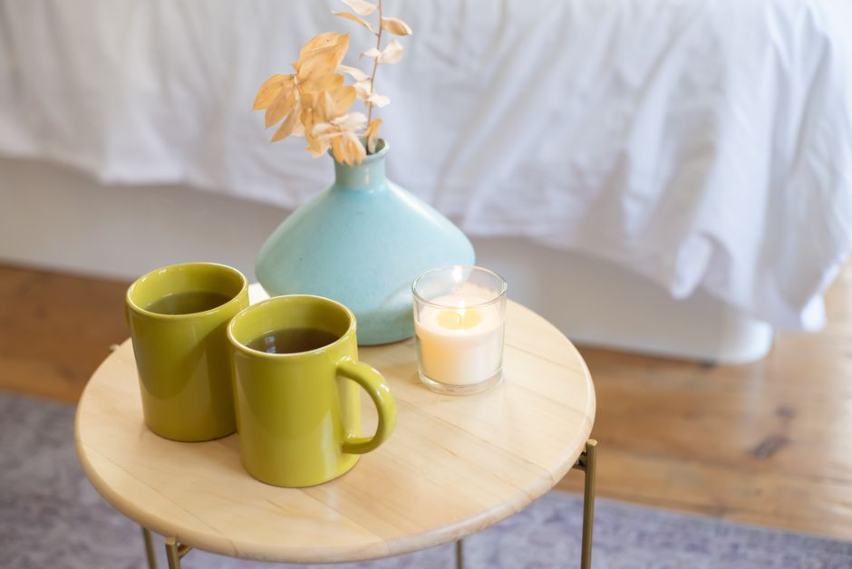 Deux tasses vertes et une bougie sur une table en bois, avec un vase bleu et des fleurs sèches en arrière-plan.