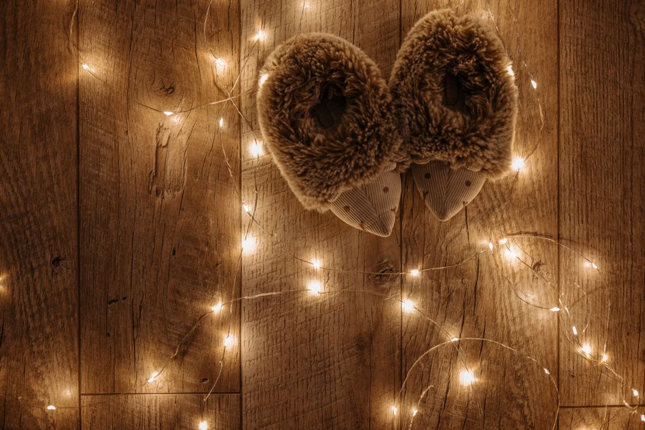 Chaussons en peluche posés sur un sol en bois entourés de guirlandes lumineuses. Ambiance chaleureuse et romantique.